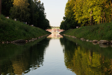 Gentle waterway lined with trees reflects a stunning palace as visitors stroll in the golden light of late afternoon.