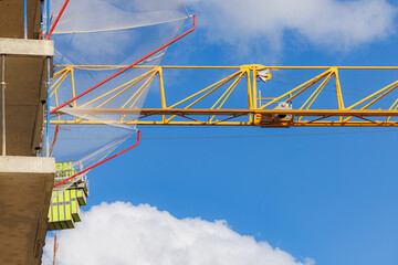A crane is lifting construction materials at a building site with fluffy clouds and a clear blue sky above.