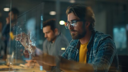 A focused man works intently at a transparent digital interface, showcasing advanced technology in a collaborative workspace, colleagues helping in teamwork