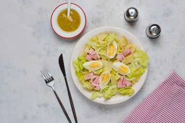Parisian salad of lettuce, boiled potatoes, ham, and hard-boiled eggs in a light-colored plate against a gray concrete background.