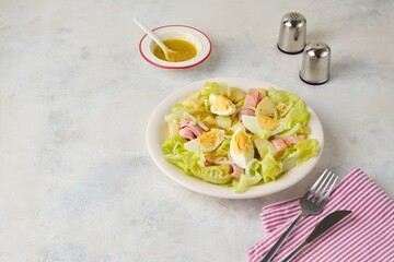 Parisian salad of lettuce, boiled potatoes, ham, and hard-boiled eggs in a light-colored plate against a gray concrete background.