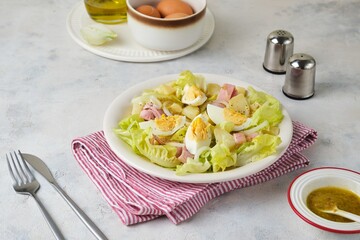 Parisian salad of lettuce, boiled potatoes, ham, and hard-boiled eggs in a light-colored plate against a gray concrete background.