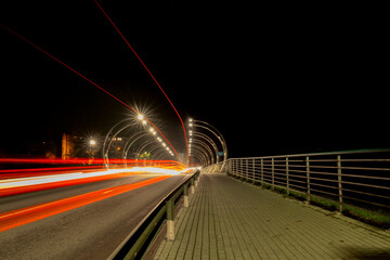 A modern bridge over the Gauja River, illuminated at night by arched street lighting fixtures and long strips of light created by passing vehicles, creates a dynamic effect.