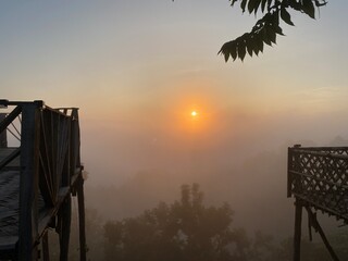 grassy mountain slope with a wooden fence, lit by morning sun. Tall evergreen overlooks hazy valleys. moody winter sunset over the Cotswold countryside. Sunlight on the morning with sea of fog.
