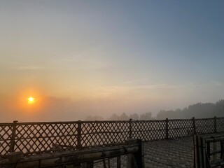 grassy mountain slope with a wooden fence, lit by morning sun. Tall evergreen overlooks hazy valleys. moody winter sunset over the Cotswold countryside. Sunlight on the morning with sea of fog.