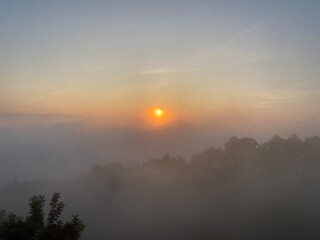 grassy mountain slope with a wooden fence, lit by morning sun. Tall evergreen overlooks hazy valleys. moody winter sunset over the Cotswold countryside. Sunlight on the morning with sea of fog.
