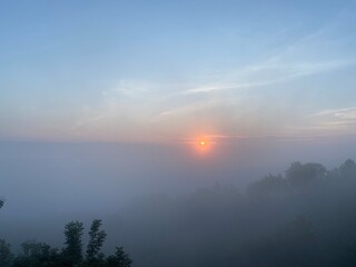 grassy mountain slope with a wooden fence, lit by morning sun. Tall evergreen overlooks hazy valleys. moody winter sunset over the Cotswold countryside. Sunlight on the morning with sea of fog.