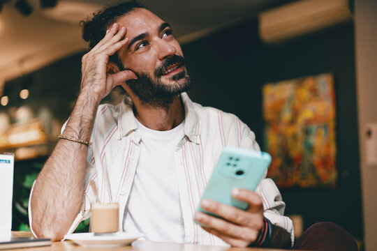 Bearded man laughing while holding smartphone at café counter with coffee. Conceptual photo about authentic joy, individuality, digital culture, and technology-driven lifestyle.