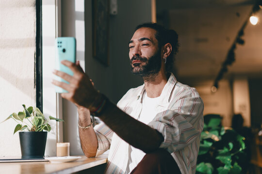 Bearded man in casual shirt looking into smartphone camera, relaxed freelancer at cafe window with coffee and laptop, representing concept of digital presence.