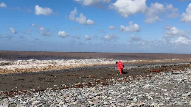 Man in bright orange suit using a metal detector on a rocky pebble beach after storm, searching for lost items or treasures along the shoreline at sunny day. Stormy waves on background
