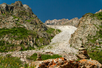 Naklejka premium Rocky mountain pass with steep slopes, scattered cairns, scree field, lush green patches, blue sky, rugged terrain, alpine environment, popular hiking and mountaineering destination, summer season