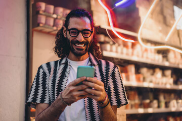 Young man laughs while using smartphone near window with neon lights, enjoying social interaction, representing joy, digital flow and modern mobile identity. © BullRun