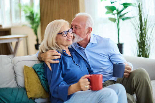 Portrait of a happy senior couple embracing talking and drinking coffee or tea at home - Powered by Adobe