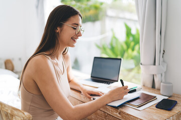 Smiling woman writes at desk, demonstrating confident workflow, focus and tech fluency in remote...