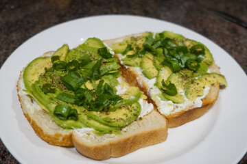 Two sandwiches with sliced avocado, Italian herbs, fresh green onions, and Philadelphia cream cheese on a large white plate.