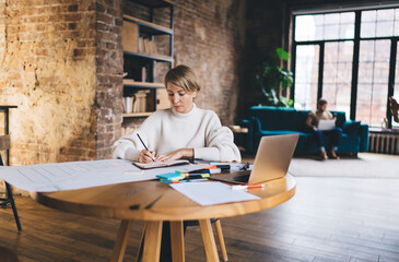 Focused young woman writes in notebook during UX planning session while male freelancer works...