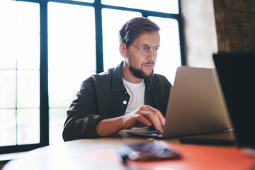 Focused male freelancer typing on laptop, developing digital project with concentration and precision, surrounded by tech essentials at modern creative workspace