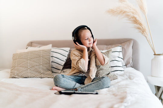 Smiling brunette mother playfully reaches for laughing daughter wearing headphones and lying with tablet, joyful family moment showing positive tech interaction and bonding.