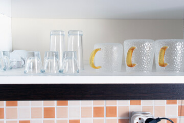 A shelf of glass glasses inside a kitchen cabinet, three outlets plugged into the wall
