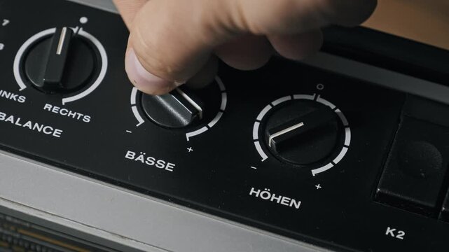 High-angle macro of a hand turning bass and treble equalizer knobs on a vintage audio system. German labels Basse and Hohen visible on the black brushed metal control panel.