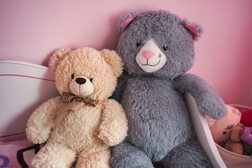Two cute teddy bears sitting together on pink bed in cozy children room interior with soft toys and warm light