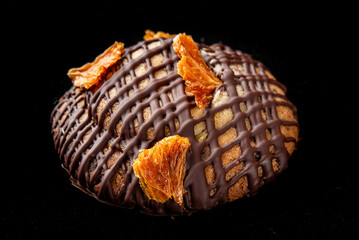Close-up of a round cookie or small cake with dark chocolate drizzle and dried orange pieces on top, isolated on black background. Gourmet dessert, styled for food photography.