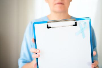 Close up of a woman doctor in medical uniform holding clipboard with blank paper and blue ribbon...