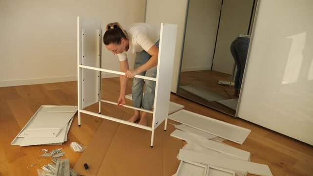 Young woman assembling white flat pack furniture at home, leaning over panel with tools and screws on wooden floor. DIY home improvement tutorial, step by step furniture setup process.