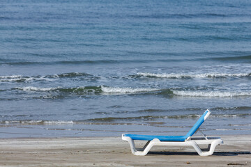 A single, empty white and blue beach lounger rests on a wide stretch of sandy shore. The empty lounger symbolizes a peaceful invitation to relaxation and off-season solitude.