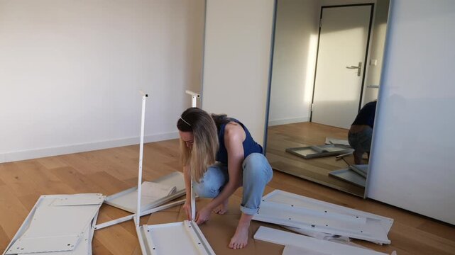Woman kneeling on wooden floor assembling white flat-pack furniture in minimal room. DIY home setup, tool in hand, mirror reflection adds depth. Real-life interior work, moving in moment.