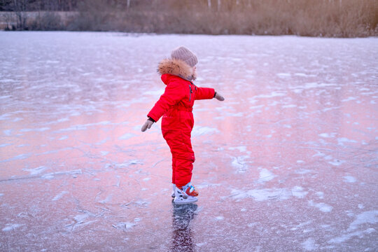 A child in a bright red jumpsuit learns to skate on the ice of a frozen lake at dusk.