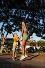 Obraz premium Joyful fit mother and her smiling little son jumping together during a workout session in the park, holding hands and laughing under the warm sunlight surrounded by trees and playground