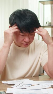 Asian man sitting at home table stares at due bills, calculator and laptop nearby, head resting in palms. Chinese guy looks worried, trying to find way to cover costs before tight payment deadline.