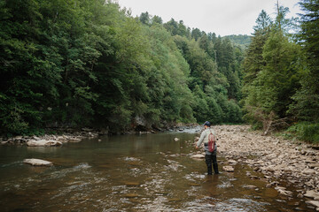 Fisherman wading in river, casting line surrounded by lush green forest