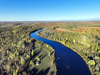 An aerial drone view captures the Indian River in Ontario as it meanders through an expansive forest landscape exhibiting vibrant autumn foliage under a clear blue sky