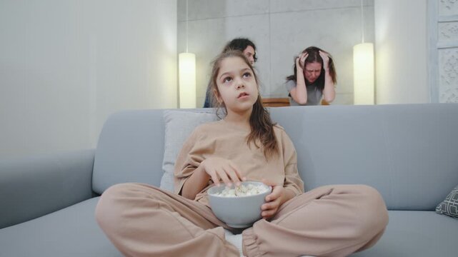A teenage girl calmly eats popcorn on the sofa, trying to tune out her parents loud fight behind her. The contrast highlights emotional detachment and tension