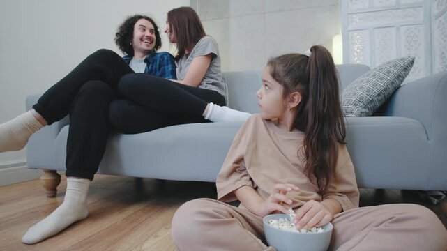 A teenage girl sits alone on the floor, visibly sad, while her cheerful parents relax and laugh on the sofa, unaware of her emotional isolation in the cozy home