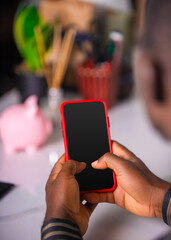Black person holding mobile phone with blank screen and a blurred background of stationary and pinky bank suggesting mobile banking, social media, news, entertainment