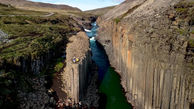 Majestic basalt columns frame the vibrant green waters of Studlagil Canyon in Iceland. Adventurers stand on rocky outcrops, appreciating the incredible landscape under a clear blue sky.