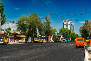Obraz premium Fresh asphalt layer being laid by paving machines and compacted by road rollers on a city street, surrounded by trees, utility wires, and residential high-rise buildings