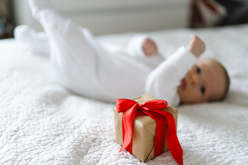 A small gift wrapped in brown paper with a red ribbon sits on a white bedspread. In the background, a baby in a white outfit is playing happily on the bed, creating a joyful atmosphere.