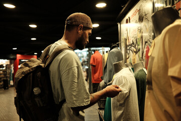 Young man with a backpack examining a T-shirt in a clothing store. Concept of retail, fashion, and lifestyle