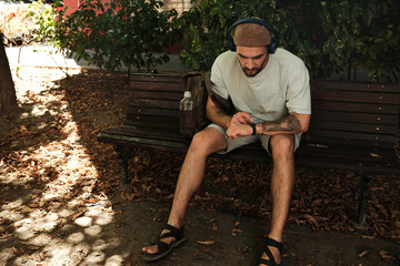 Young man sitting on a park bench with headphones, checking smartwatch. Concept of relaxation, health tracking, and lifestyle