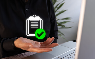A businessman, sitting at his desk, is holding a clipboard with a green check mark on it.
