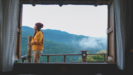 A woman standing on wooden balcony with mountain views on foggy morning outside the window
