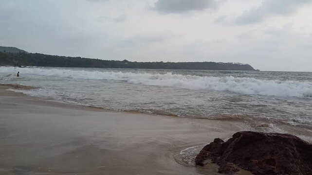 Powerful ocean waves crashing onto a sandy shore near a rocky outcrop, with a distant, forested headland under a soft, overcast sky. Ideal for projects related to nature, travel, environment.