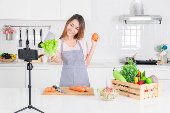 Smiling Asian woman in an apron hold lettuce and a bell pepper while recording a cooking tutorial video in her bright white kitchen with a smartphone on a tripod.