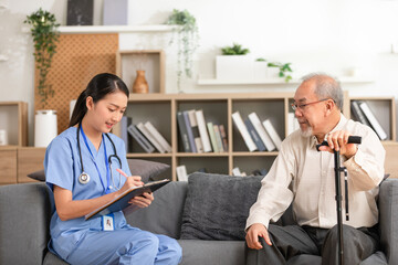 Asian Young nurse in blue scrubs speaking with elderly man with cane on living room sofa, caring medical home visit and friendly conversation