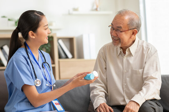 Asian friendly nurse in blue scrubs handing medication bottle to smiling elderly man at home consultation with caring expression and warm lighting