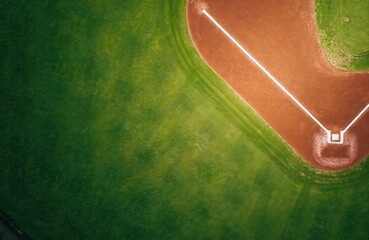 Aerial view depicts a baseball diamond detail. Green grass covers outfield area. Dirt infield displays base lines and home plate. This organized sport theme shows recreation ground and team venue.